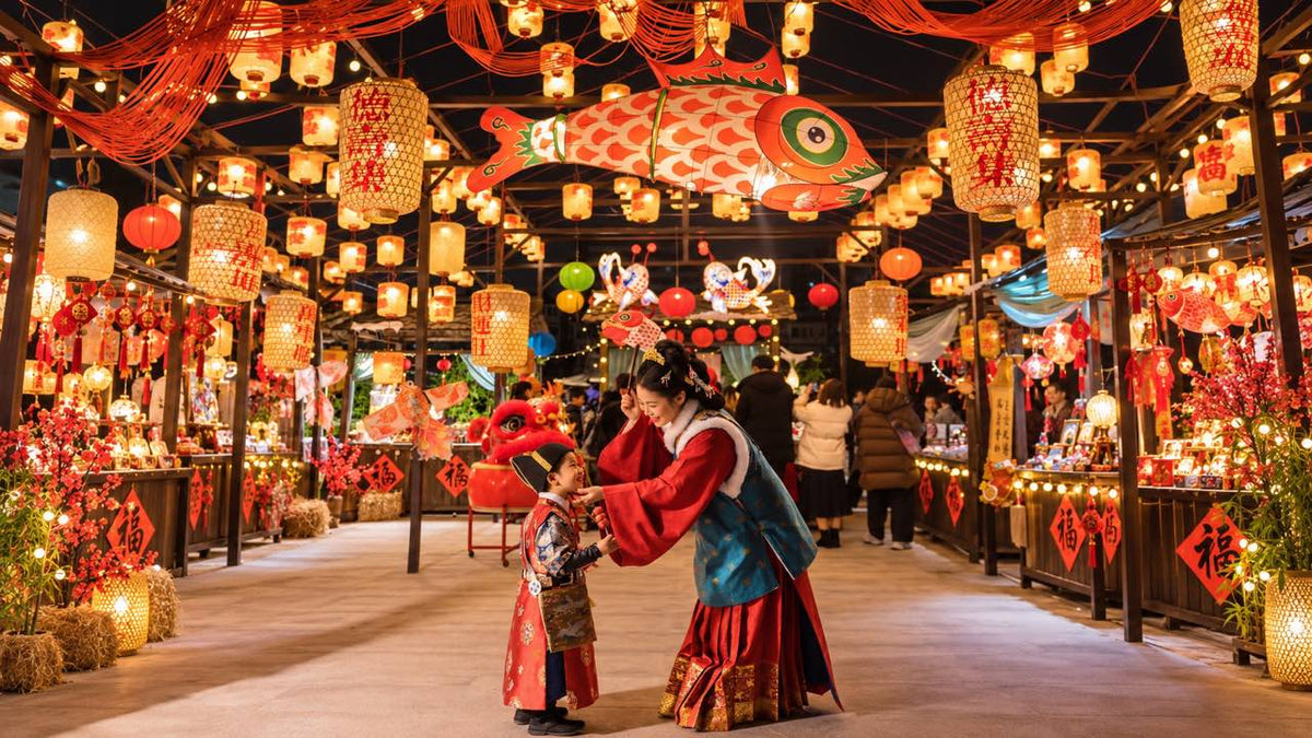 A mother and her young son in hanfu smiling at each other beneath glowing fish lanterns at a Chinese New Year market.