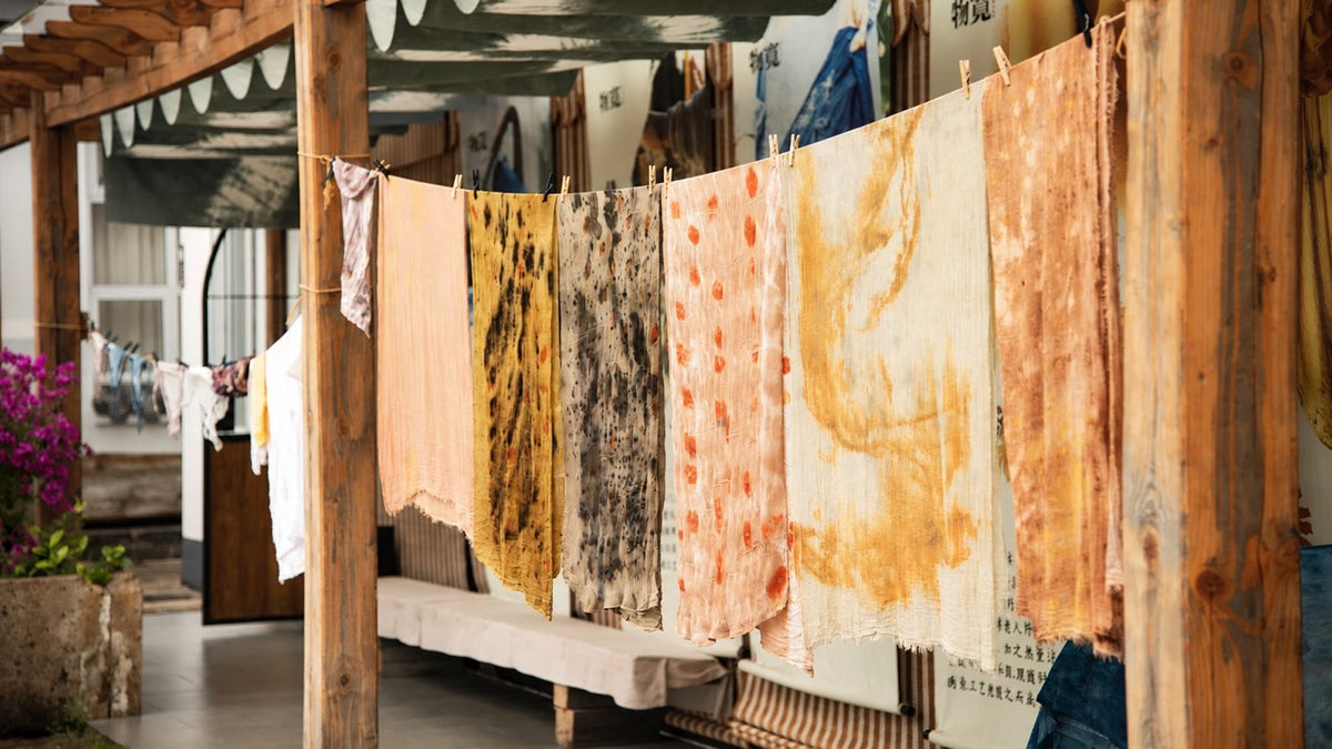 Plant-dyed fabrics in yellow, peach and gray hanging to dry under a wooden veranda.